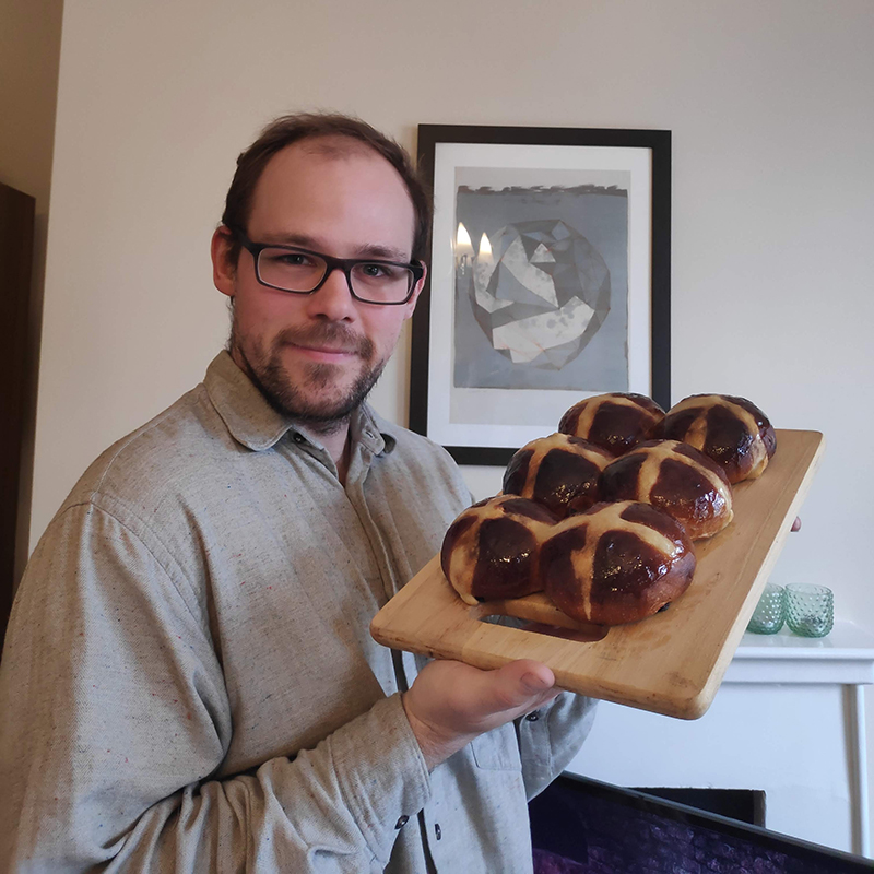 A photo of baker Ben proudly holding a wooden hopping board laden with hot cross buns. There is a piece of abstract art on the wall in the background of the image