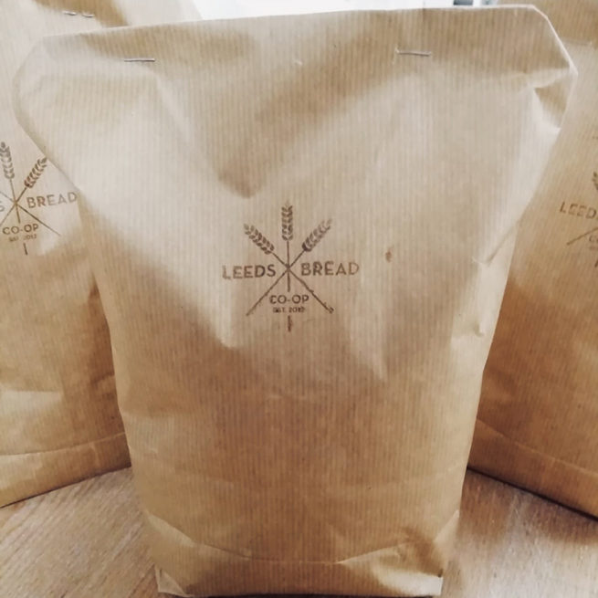 A photo of a bag of flour, the bag is brown paper with the Leeds Bread Co-op logo stamped on it in brown ink