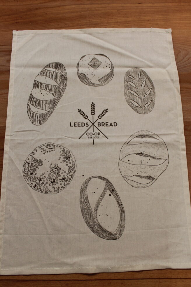 a cream-coloured tea towel, the the middle is the Leeds Bread Co-op logo in brown, and surrounded by a circle of hand illustrated loaves, also in brown ink.