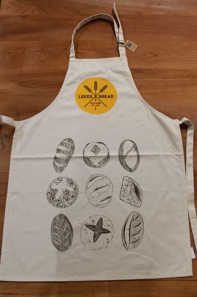 A cream coloured apron with an adjustable neck strap. At the top of the apron is the Leeds Bread Co-op logo, brown text in a yellow circle. Below that are nine hand illustrated loaves in a 3x3 grid.
